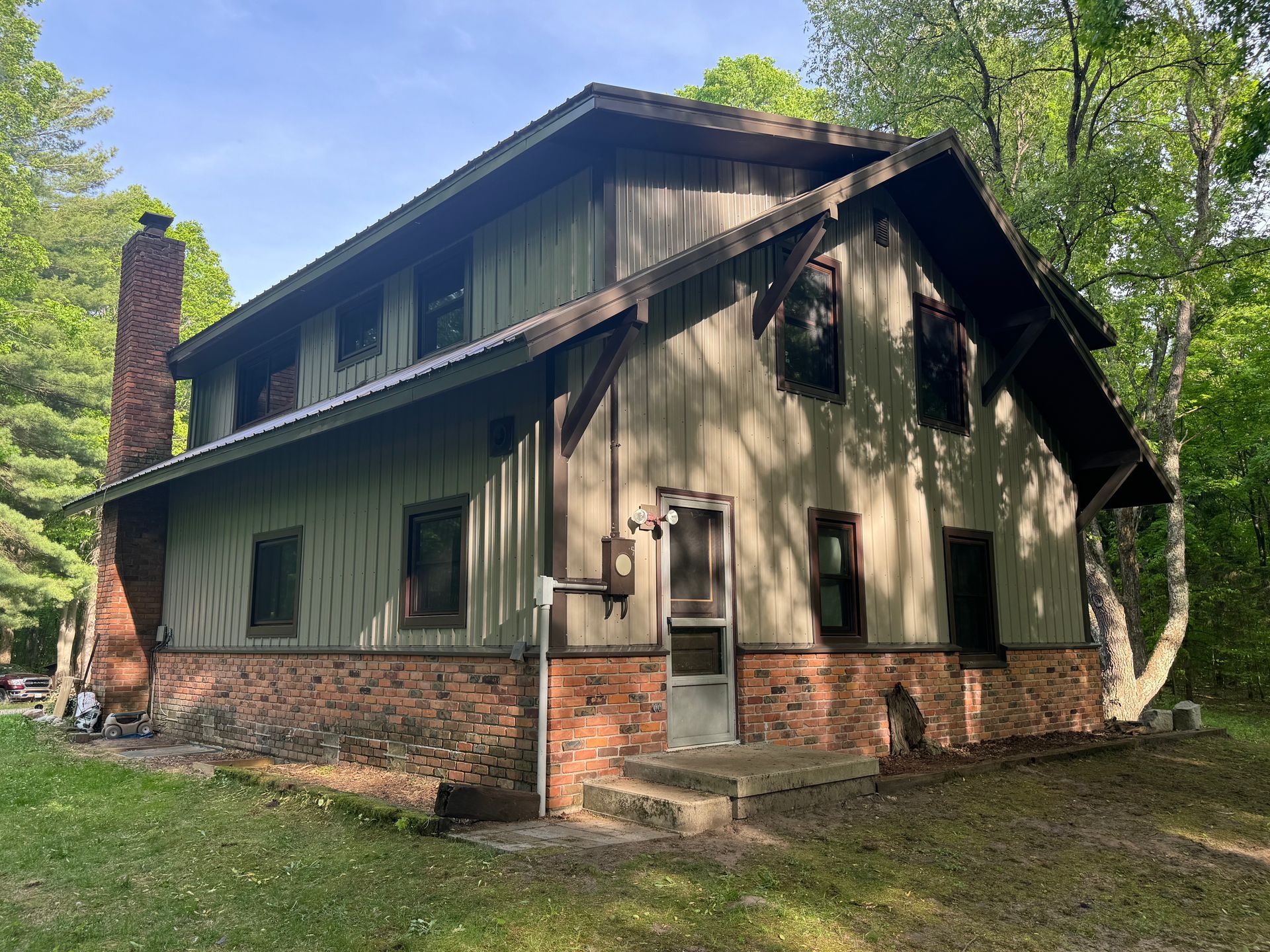 Two-story rustic cabin with brick base, dark green siding, and a tall chimney among trees