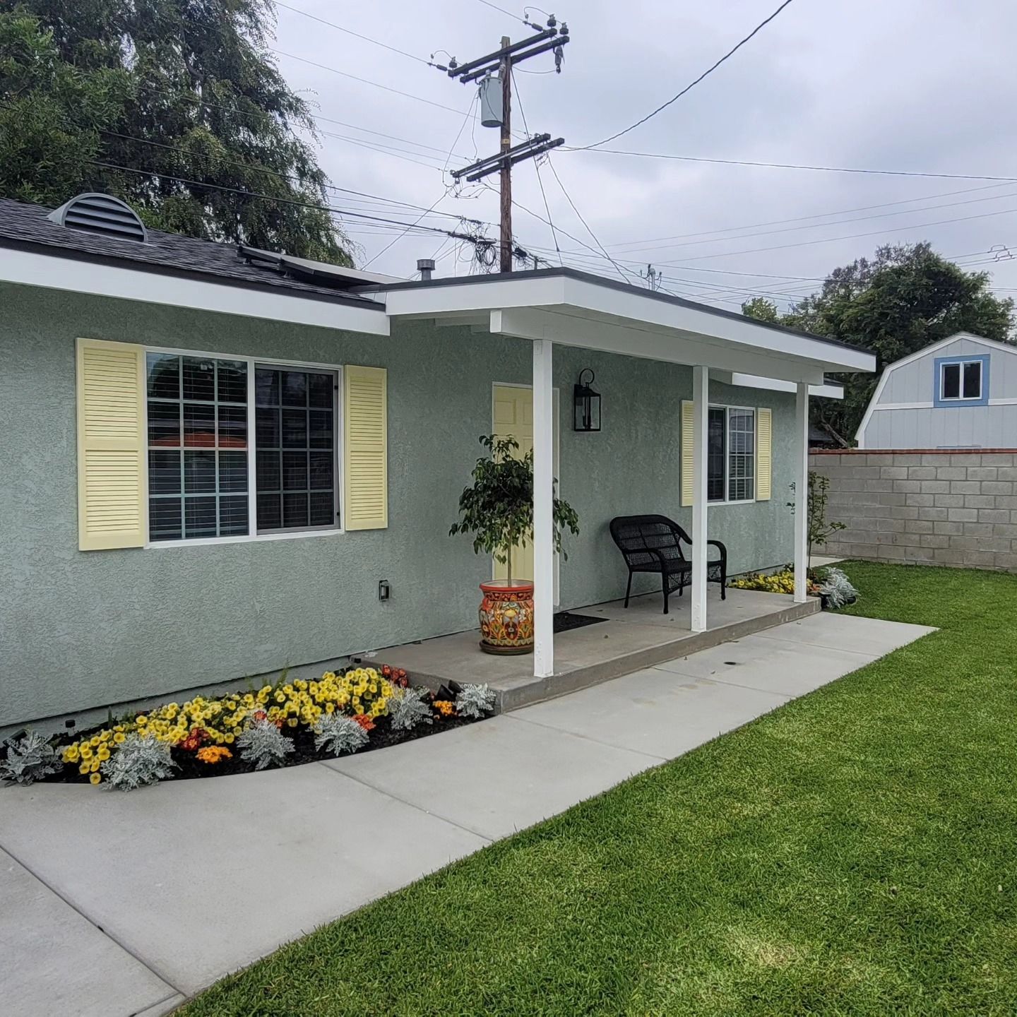 Small gray house with yellow shutters, flower bed, porch chair, and green lawn under cloudy sky