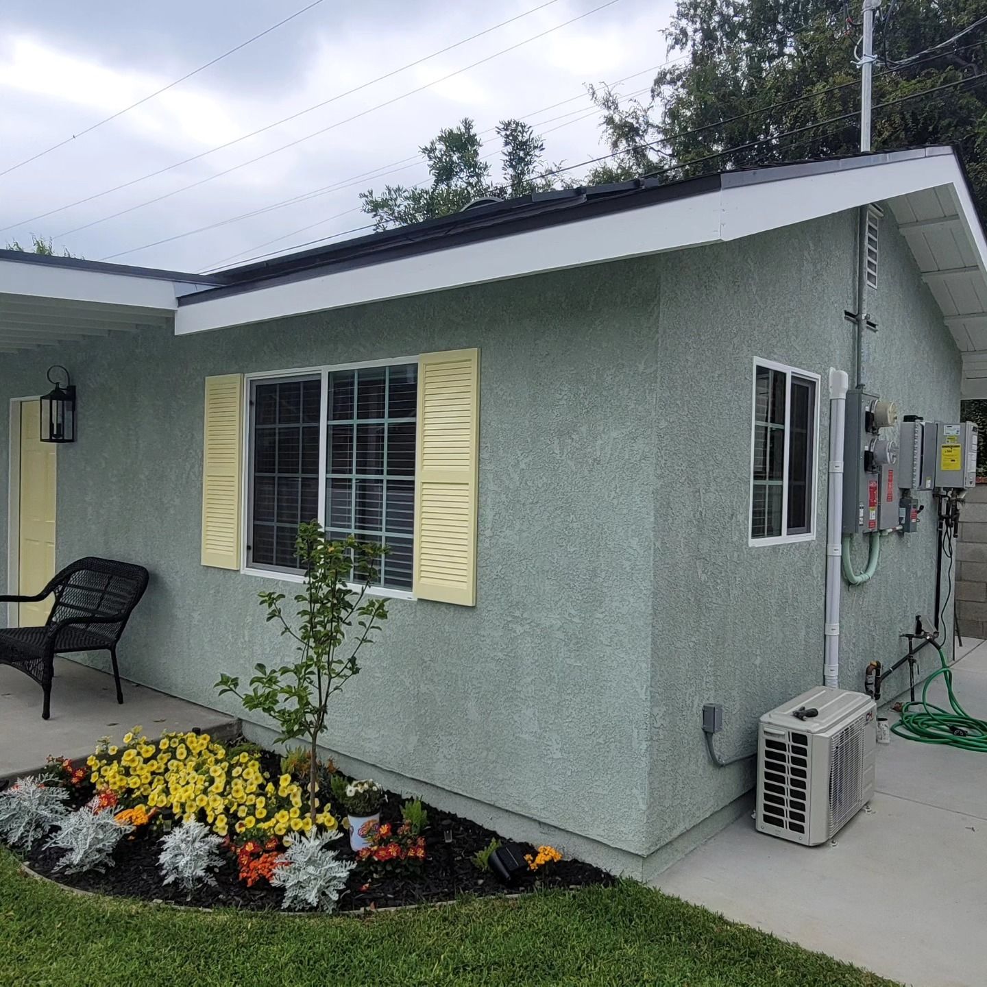 Small stucco house exterior with flower bed, yellow shutters, and air conditioner unit beside a patio