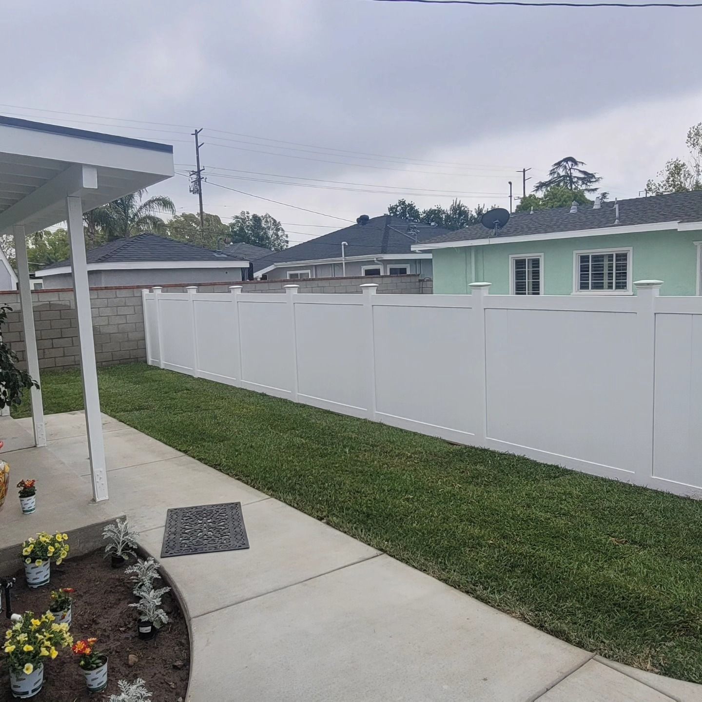 Backyard patio with concrete walkway, flower bed, green lawn, and white privacy fence beside a mint house