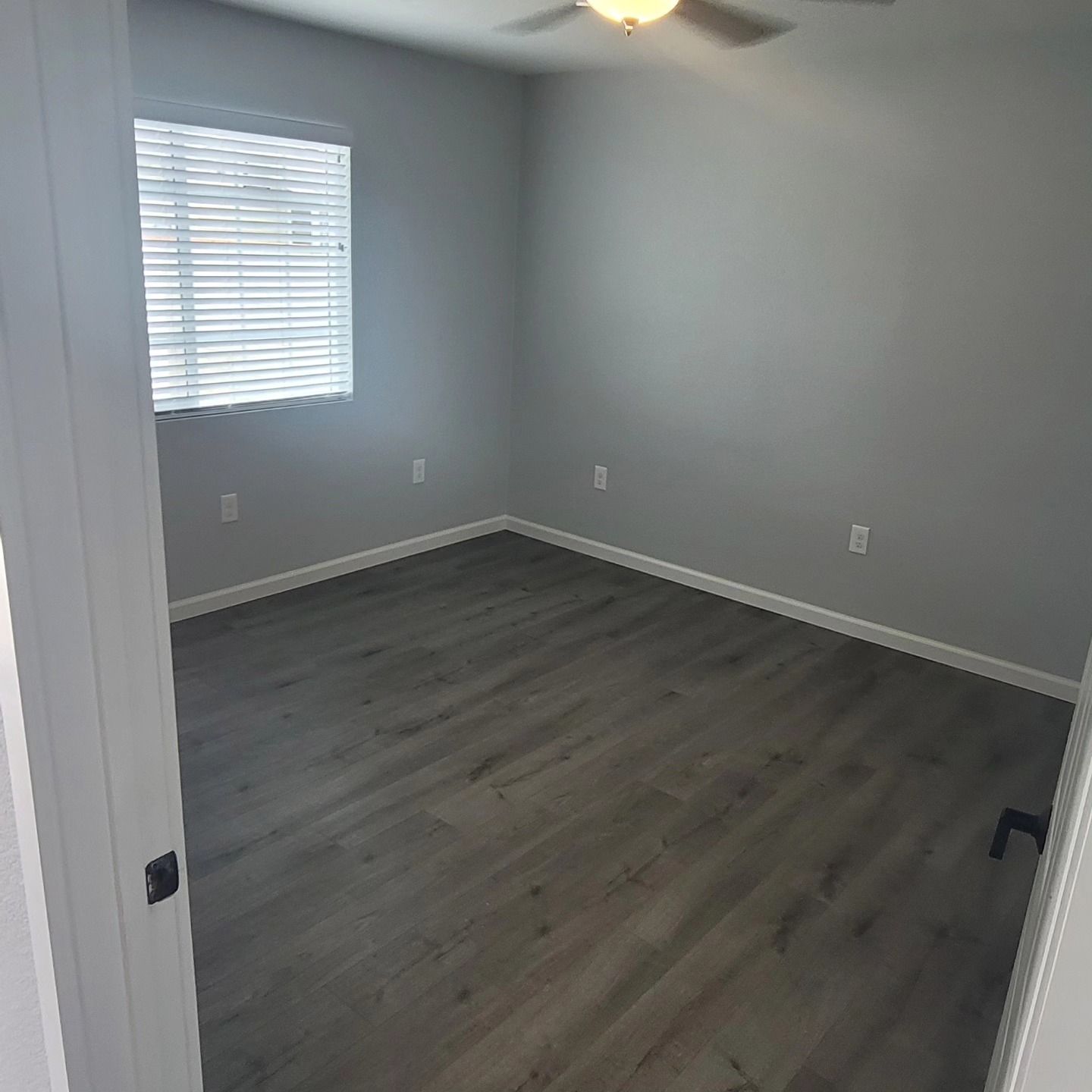 Empty gray bedroom with a window, white trim, and dark wood-style flooring.