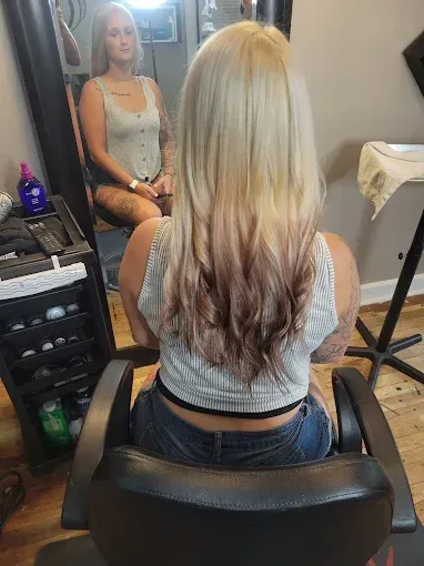 A person sits in a salon chair with long blonde hair featuring a rose-gold ombre effect, viewed from behind in a mirror.