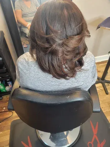A person with shoulder-length, dark brown hair with soft, layered curls sits in a salon chair facing a mirror.