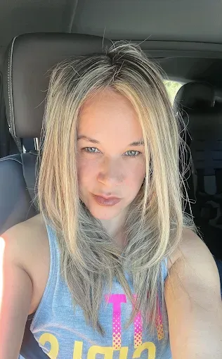 A person with blonde, shoulder-length hair wearing a grey graphic tank top takes a selfie inside a vehicle.