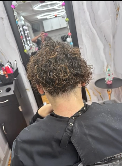 A person with short, curly, highlighted hair sits in a salon chair with a cape, facing a mirror.