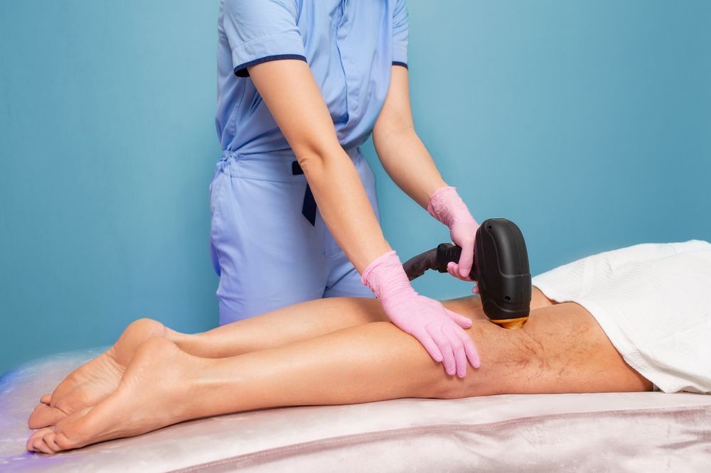 A technician in blue scrubs and pink gloves uses a laser hair removal device on a person’s leg.