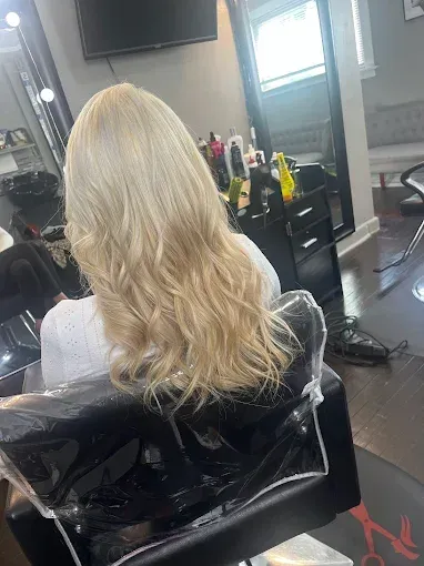 A person with shoulder-length, wavy blonde hair sits in a salon chair, seen from behind.
