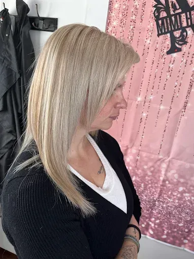 Profile view of a person with shoulder-length, straight blonde hair styled in a salon, standing against a pink backdrop.