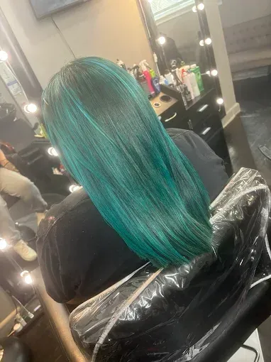 A person with long, straight, teal-dyed hair sitting in a salon chair facing a vanity mirror.
