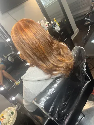 A person with shoulder-length, light brown hair with blonde highlights sits in a salon chair facing a mirror.