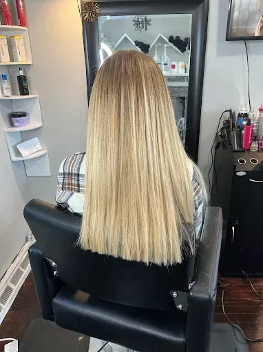A person with long, straight, blonde hair sits in a salon chair, facing a mirror.