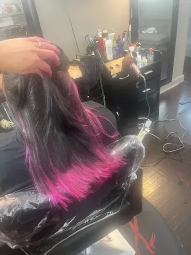 A stylist's hand separates dark hair to reveal vibrant magenta-pink colored tips in a salon setting.