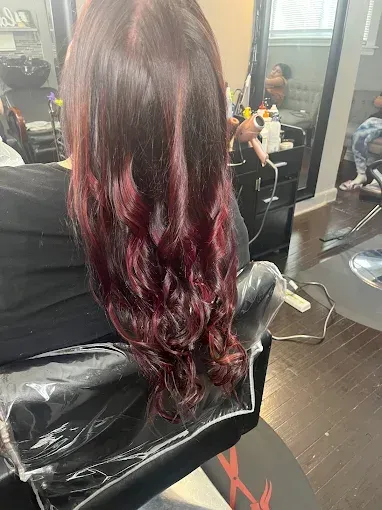 A person with long, dark auburn-toned hair with loose curls, seen from behind in a hair salon setting.