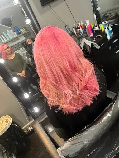 A person with vibrant pink, shoulder-length wavy hair sits in a salon chair, seen from behind with a mirror reflection.