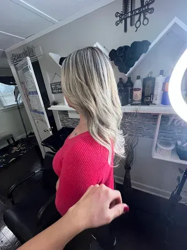 A person with shoulder-length blonde wavy hair sits in a salon chair, seen from behind with a hand reaching toward them.