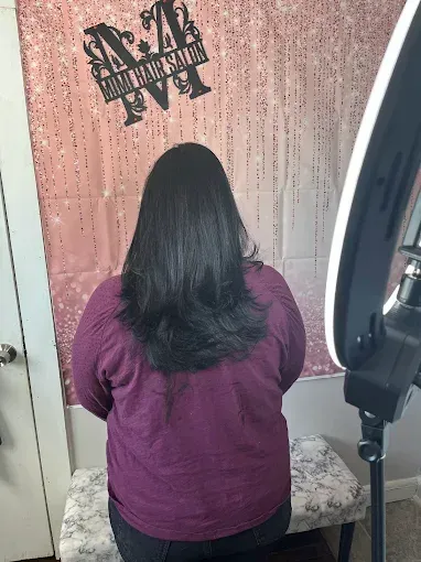 A person with dark, layered hair seen from behind in a salon, next to a ring light and a wall sign.