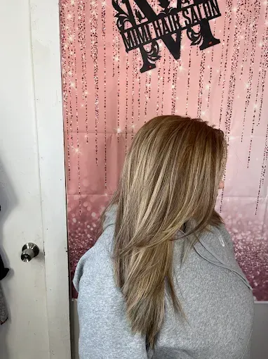 A person with long, layered, honey-blonde highlighted hair seen from the back in a salon with a pink backdrop.