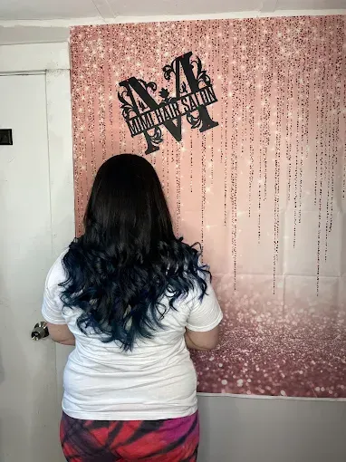 A person with long, wavy dark hair with blue-toned ends stands in front of a rose-gold glitter backdrop with a salon logo.
