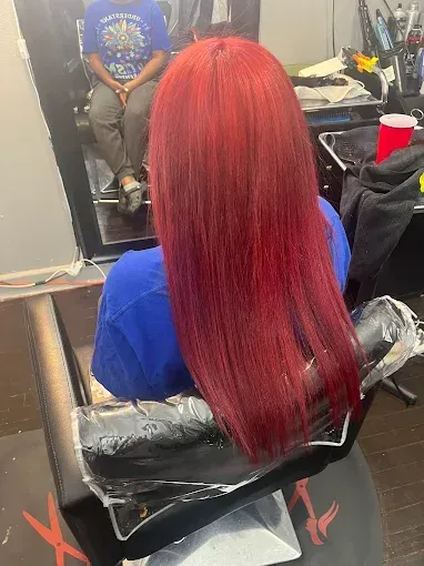 A person with bright red hair sits in a salon chair, seen from behind, with their reflection visible in a mirror.