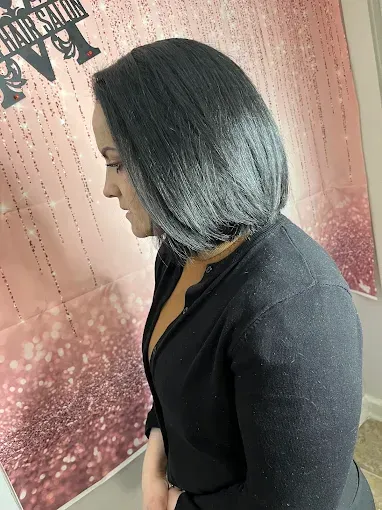 A person with shoulder-length, dark gray ombre hair stands against a pink, glittery backdrop in a salon.
