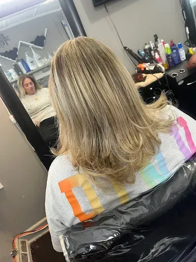 A person with shoulder-length, blonde-highlighted hair sits in a salon chair, reflected in a mirror.