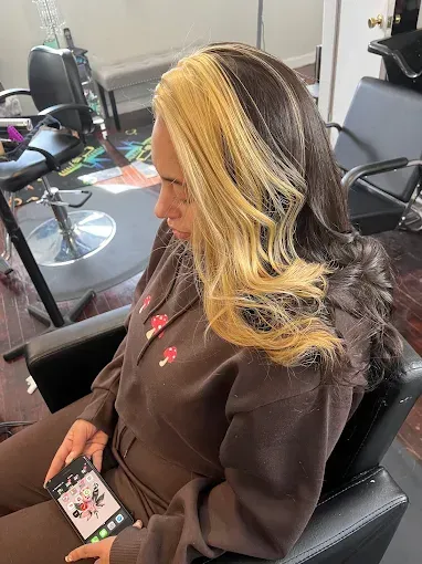 A person with long, two-toned hair—dark brown and bright blonde—sitting in a salon chair while looking at a smartphone.