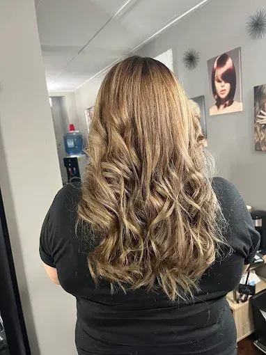 A view from behind of a person with long, wavy, highlighted light brown hair in a salon setting.