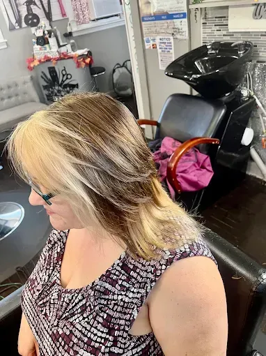 A person with two-toned brown and blonde hair, styled with layers, sits in a salon chair with a wash basin in the background.