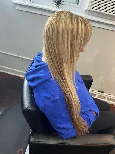 A person with long, straight, highlighted blonde hair sits in a salon chair, wearing a blue hoodie.