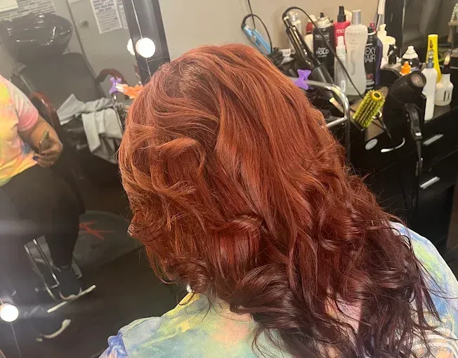 A close-up of wavy, vibrant copper-red hair in a salon setting with styling tools visible in the background.