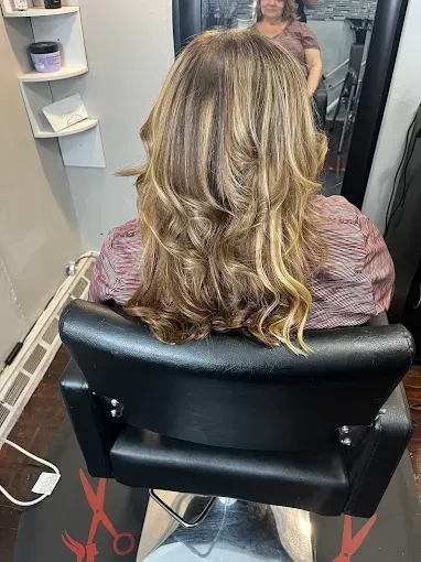 A person with highlighted, shoulder-length wavy brown hair sits in a salon chair facing a mirror.