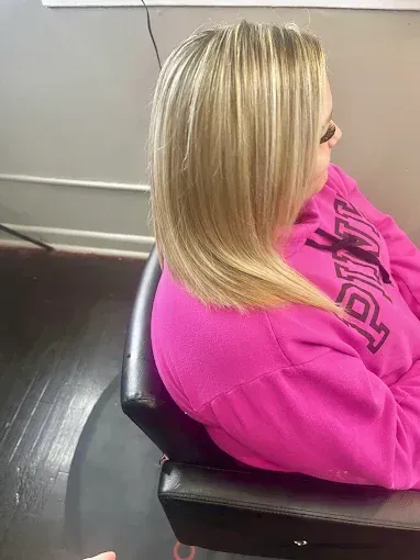 A person with shoulder-length blonde highlighted hair sits in a salon chair, wearing a bright pink sweatshirt.