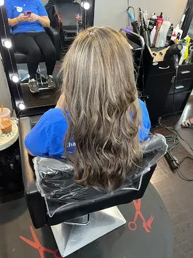 A person with long, wavy, highlighted brown hair sits in a salon chair facing a mirror, wearing a blue shirt.