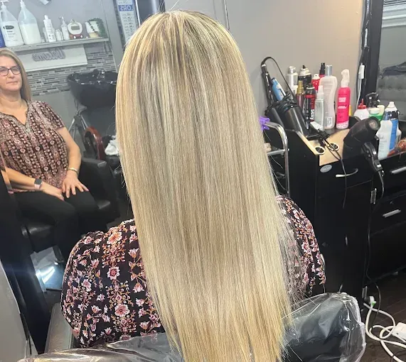A person with long, straight blonde hair with highlights sits in a salon chair, seen from behind.