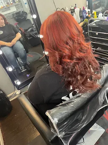 A person in a salon chair with vibrant, shoulder-length red hair, viewed from behind with their reflection in a mirror.