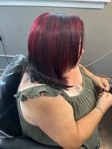 A person wearing a green top sitting in a salon chair, showing their hair with highlights of deep red and black.