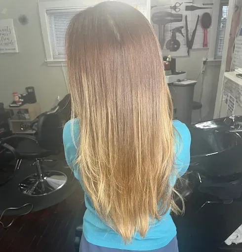 A person with long, straight, layered hair in a light brown to blonde ombre style, seen from behind in a salon setting.