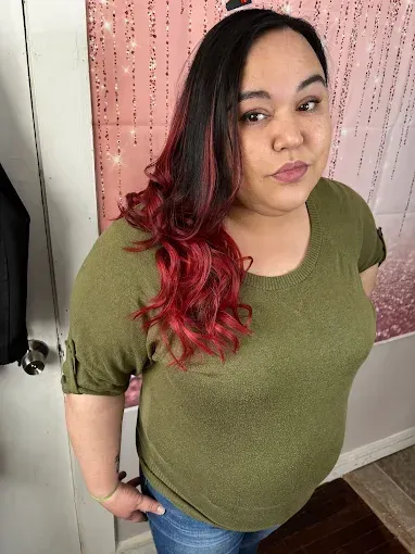 A person with dark hair transitioning to vibrant red waves, wearing an olive green shirt in front of a pink backdrop.