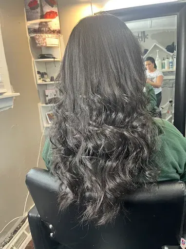 A view from behind of long, wavy dark brown hair styled in a salon chair, with a stylist visible in a mirror reflection.