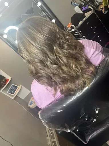 A person with shoulder-length, wavy brown hair with highlights, sitting in a salon chair in front of a mirror.