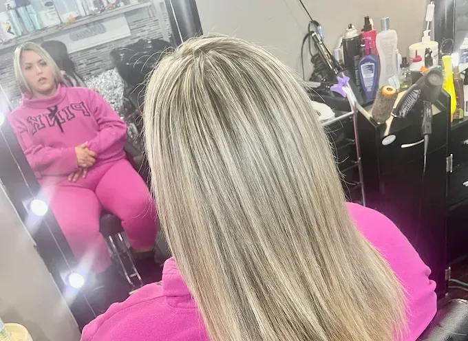 A view from behind of a person with blonde highlighted hair in a salon, seen in a mirror while wearing a pink tracksuit.