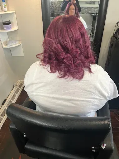 Back view of a person in a salon chair with wavy, dark magenta hair, visible in the mirror reflection.