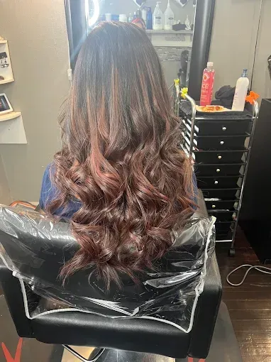 A person with long, wavy, dark brown hair featuring subtle reddish-brown highlights, sitting in a salon chair.