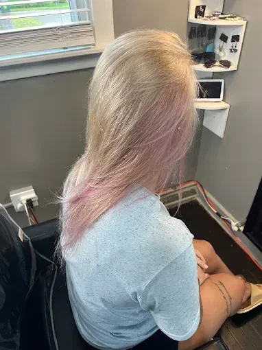 A person with platinum blonde hair with soft pink tips sits in a salon chair with a display shelf in the background.