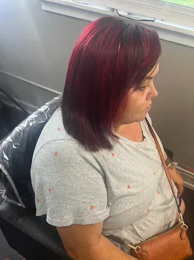 A person sits in a chair with shoulder-length, vibrant magenta-red hair, wearing a gray patterned t-shirt.