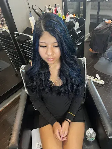 A person sits in a salon chair with long, wavy, dark blue hair, wearing a black long-sleeved dress.