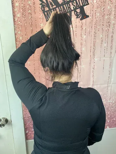 A person with dark hair wears a black long-sleeved shirt while holding a ponytail up in front of a salon backdrop.