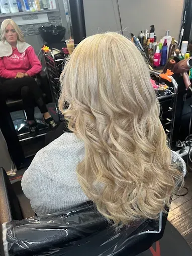 A person with long, blonde, wavy hair sits in a salon chair, facing a mirror in which another person is visible.