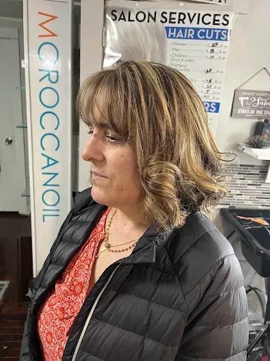 A person with shoulder-length, highlighted wavy hair and bangs stands in a hair salon.
