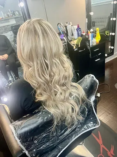 Long, wavy, blonde hair styled in a salon, viewed from behind with a mirror and styling products in the background.